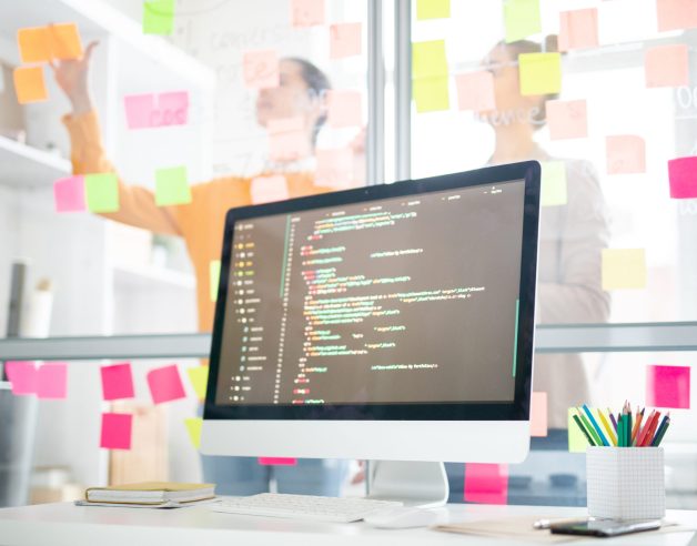 Monitor of computer with coded data and office managers planning work and sticking notes on board on background